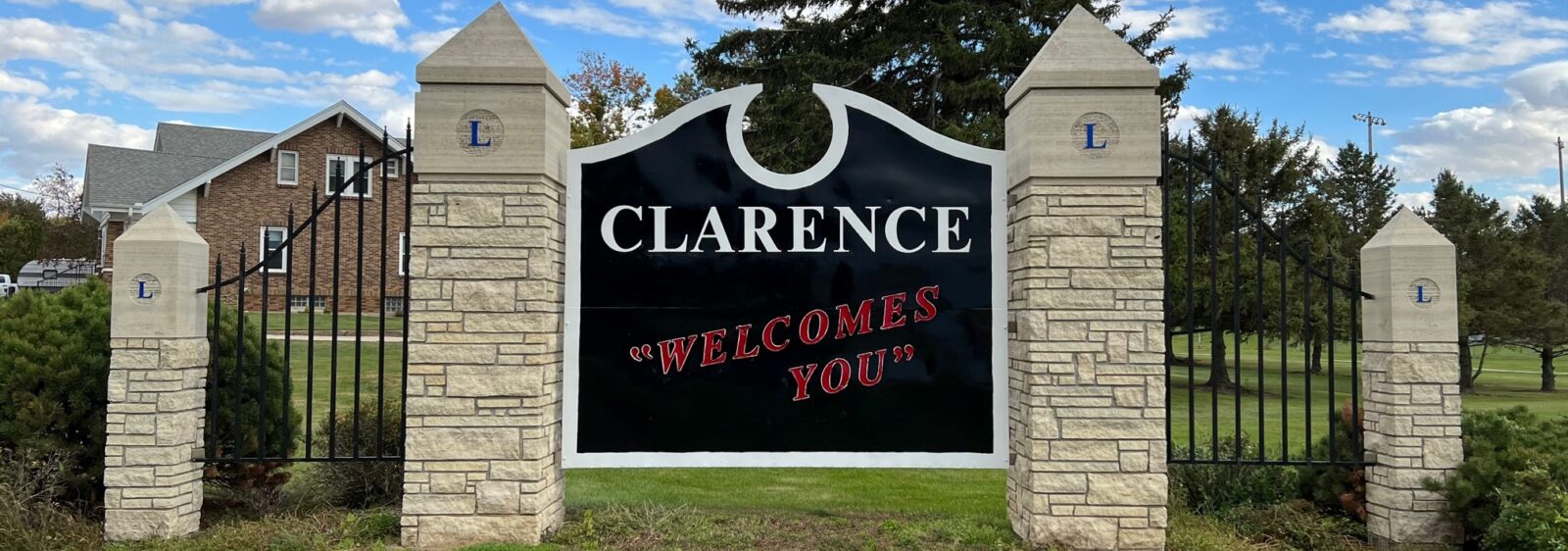 Large repainted community entrance sign reading 'Clarence Welcomes You' set in a landscaped lawn along the roadside.